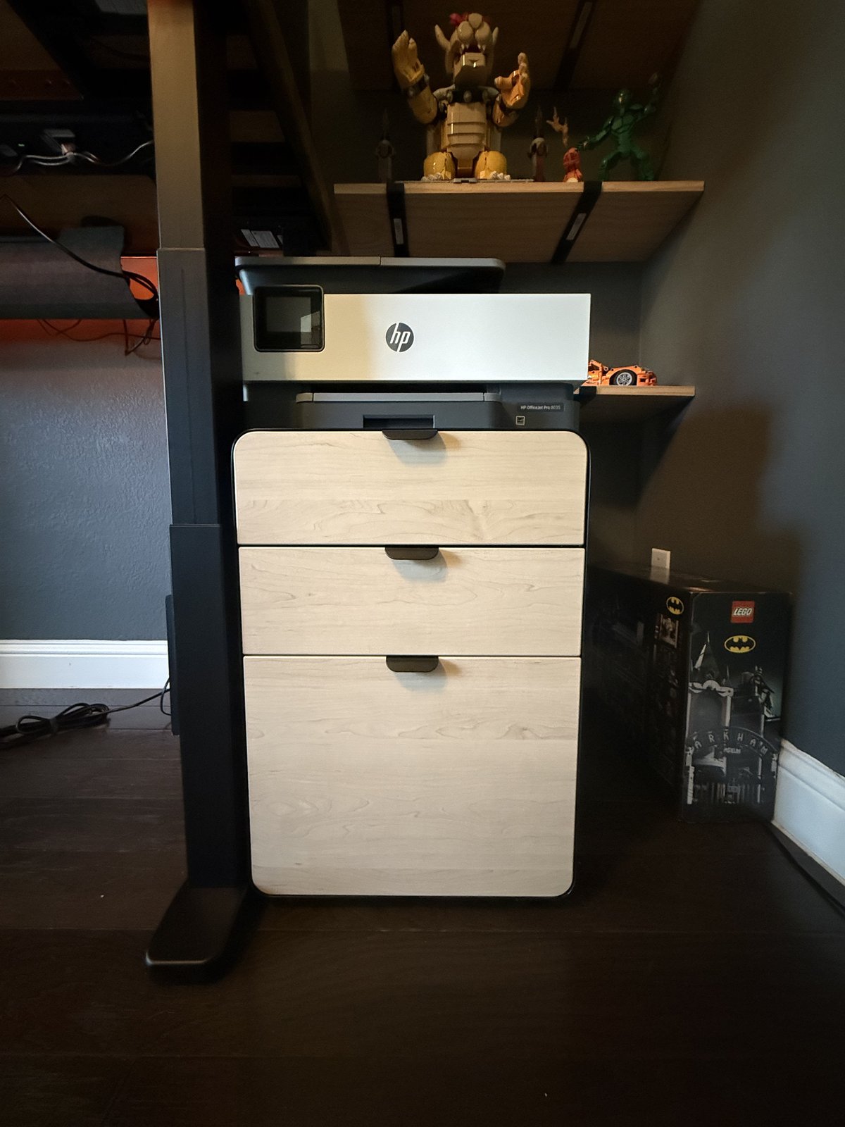The UPLIFT 3-Drawer File Cabinet with light wood drawer fronts and black pulls, an HP printer on top, beside the desk leg